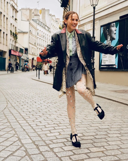 A smiling young woman in a black leather jacket poses on a cobblestone street in Paris, France, wearing patterned tights and Mary Jane shoes.