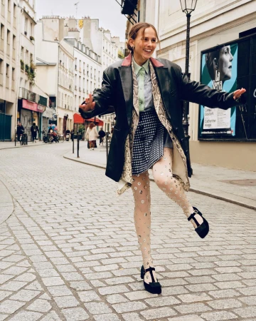 A smiling young woman in a black leather jacket poses on a cobblestone street in Paris, France, wearing patterned tights and Mary Jane shoes.