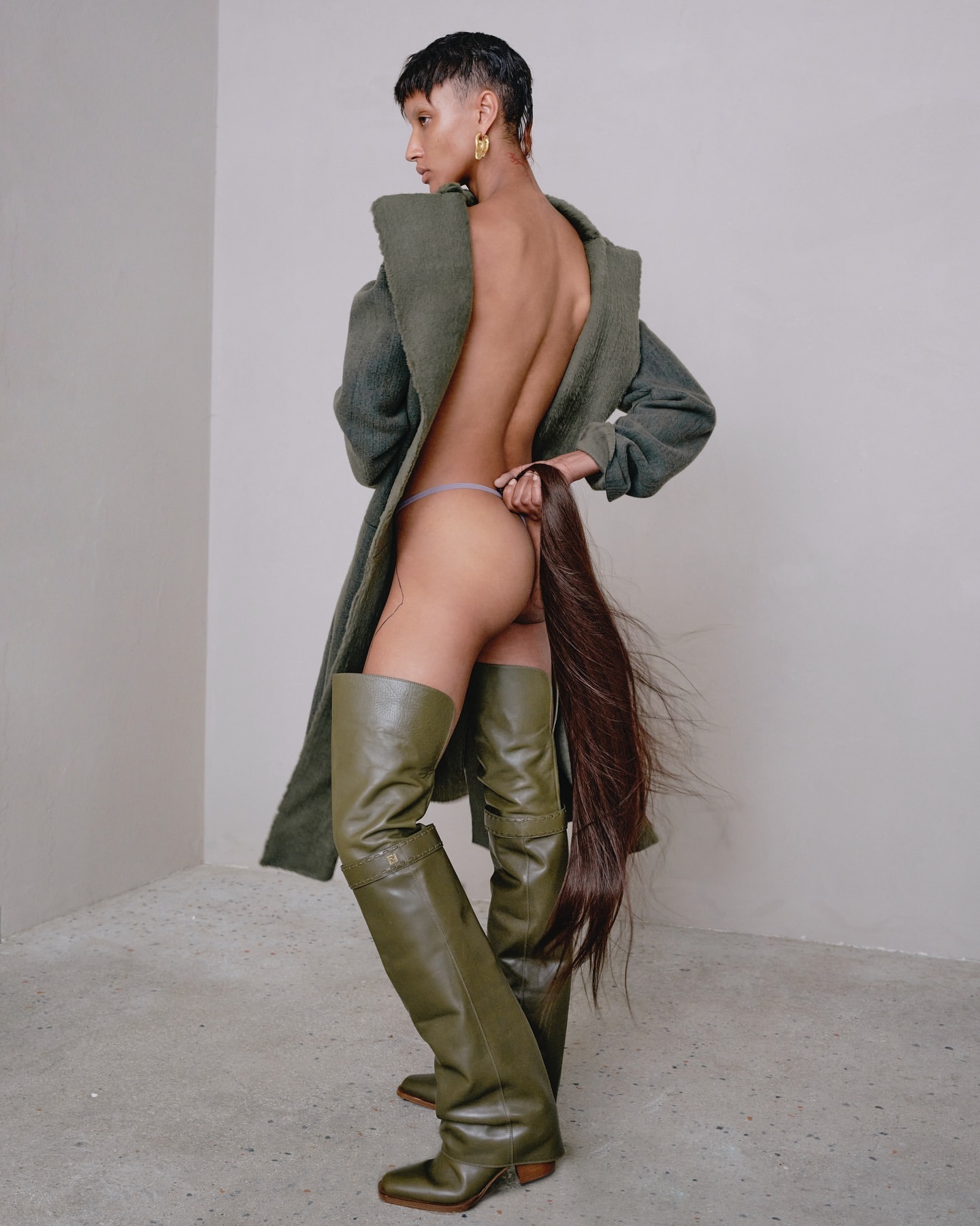 Woman in thigh-high olive-green boots and fuzzy sage coat poses in a corner studio holding her ponytail. Beauty and fashion shot.