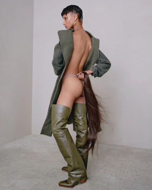 Woman in thigh-high olive-green boots and fuzzy sage coat poses in a corner studio holding her ponytail. Beauty and fashion shot.