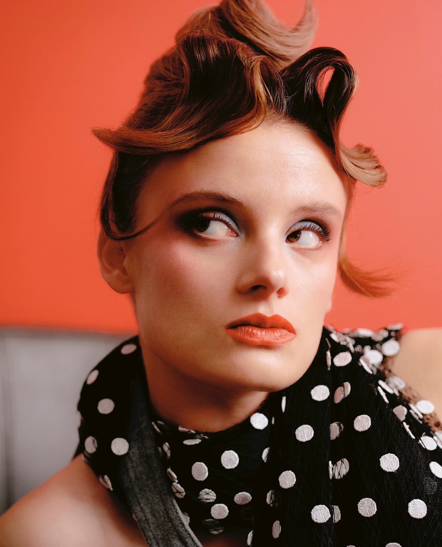 Close-up portrait of a woman with styled brown hair, striking makeup, and a polka dot scarf against an orange backdrop in a studio shot.