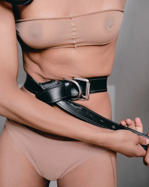 Close-up of an adult female wearing nude-color undergarments and a wide, black leather belt with a large metal buckle, holding the belt.