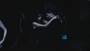Man and woman dancing in a dimly lit indoor setting, embracing closely and dressed in dark clothing, creating an intimate atmosphere.