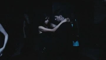 Man and woman dancing in a dimly lit indoor setting, embracing closely and dressed in dark clothing, creating an intimate atmosphere.