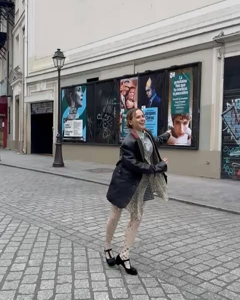 A woman in a black coat stands on a cobblestone street in front of several movie posters in an urban environment during daytime.