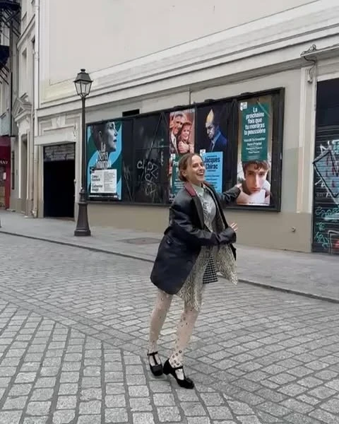A woman in a black coat stands on a cobblestone street in front of several movie posters in an urban environment during daytime.