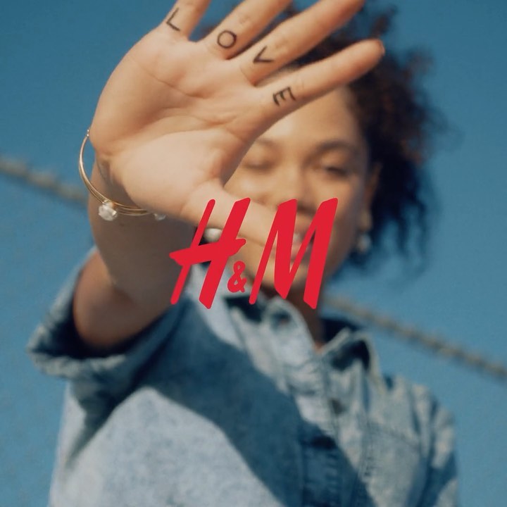 Fashion shot featuring a young adult with 'LOVE' written on their hand in front of a blurred H&M logo and light blue background.