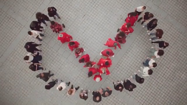 Aerial view of a large group of people forming the shape of a check mark on an outdoor paved surface. The people are wearing different colored clothes.