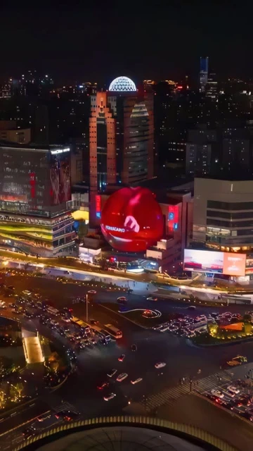 High-angle nighttime view of city intersection with vehicle traffic and illuminated buildings, including one with a large sphere.