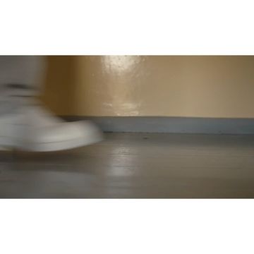 Blurred image of a person wearing a white sneaker, walking on a gray floor in a neutral-toned room with a tan wall in the background.