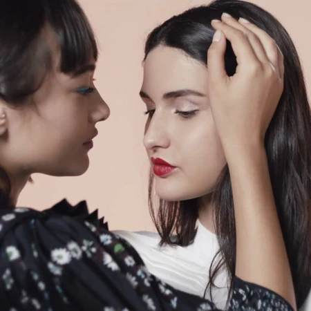Two women posing closely with one hand gently touching her hair, captured in soft studio lighting against a neutral backdrop by Alice Rosati.