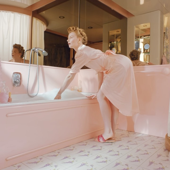 Woman in pink robe prepares bath in pink-tiled bathroom with lavender accents, reflected in mirror above tub, indoor setting.