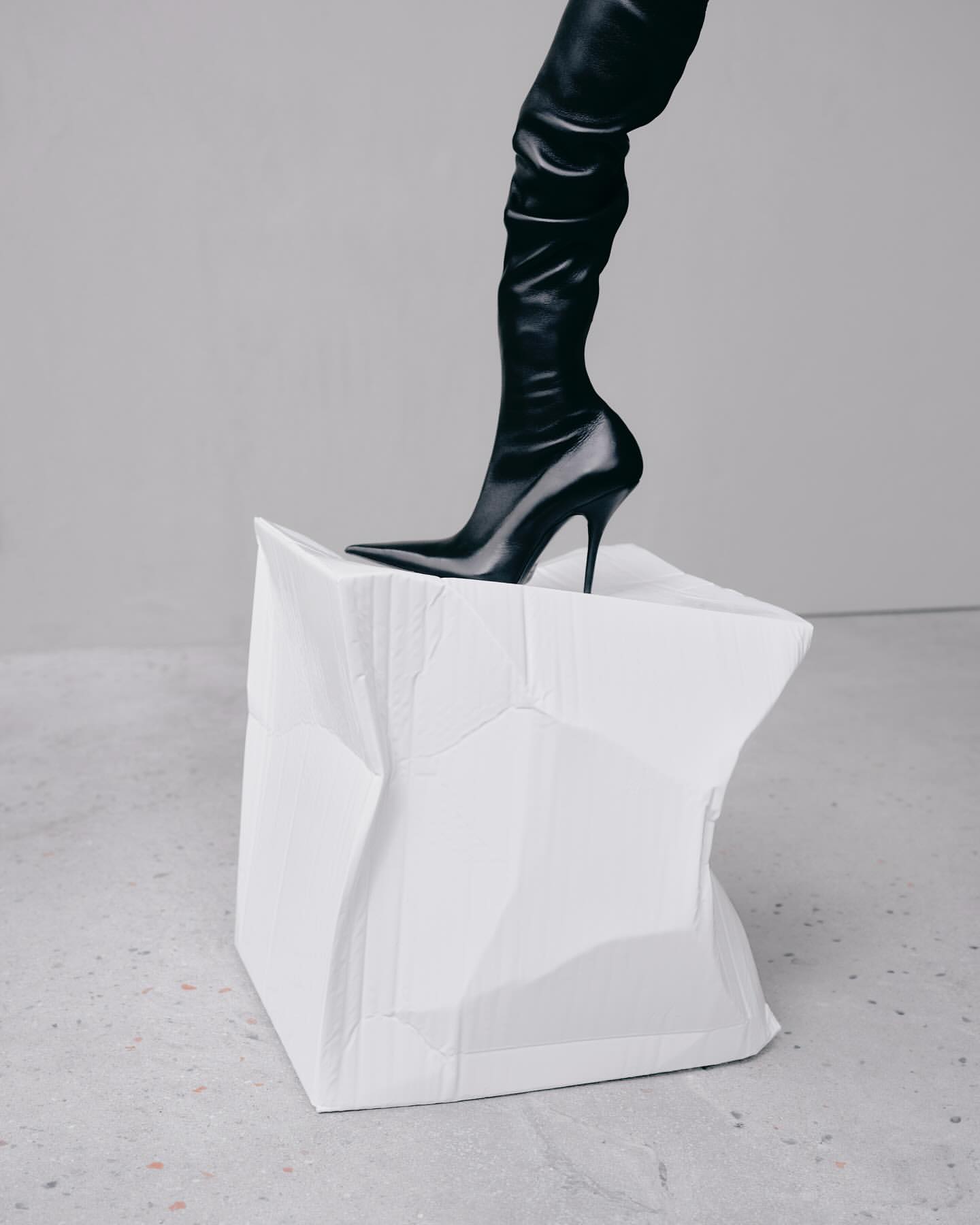 A black thigh-high stiletto boot is stepping onto a crushed white paper cube against a light grey backdrop in a studio-style shot.