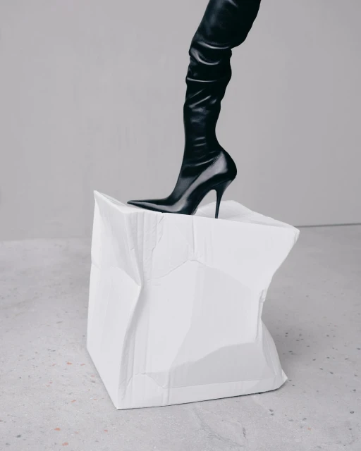 A black thigh-high stiletto boot is stepping onto a crushed white paper cube against a light grey backdrop in a studio-style shot.