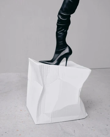 A black thigh-high stiletto boot is stepping onto a crushed white paper cube against a light grey backdrop in a studio-style shot.