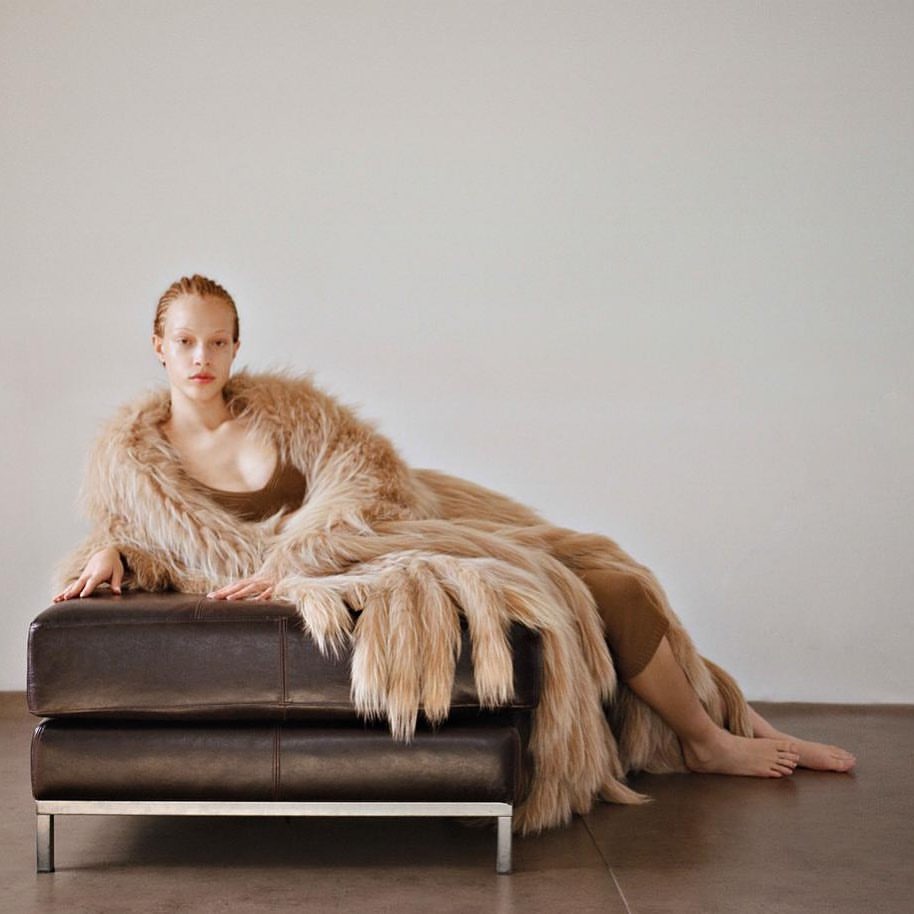 Elegant woman in a faux fur coat reclining on a leather ottoman in a light studio setting, creating a high fashion editorial image.