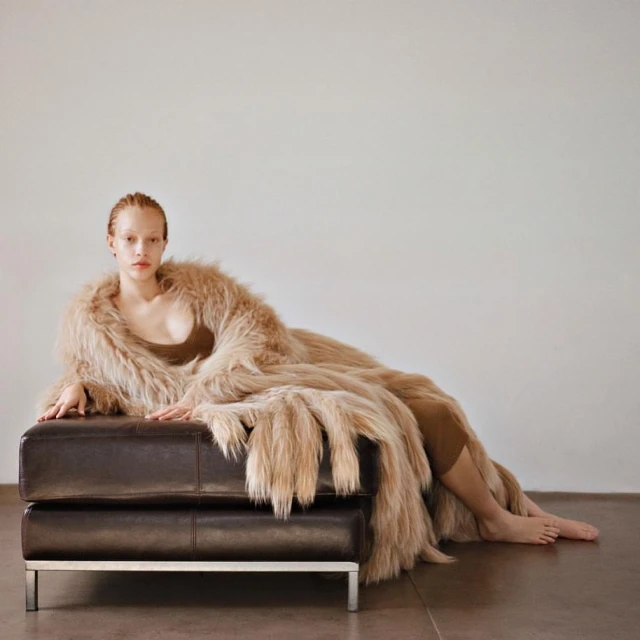 Elegant woman in a faux fur coat reclining on a leather ottoman in a light studio setting, creating a high fashion editorial image.