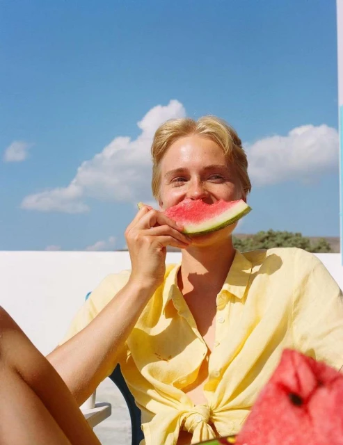 A woman with blond hair smiles while holding a slice of watermelon to her face outdoors on a sunny day with a blue sky.