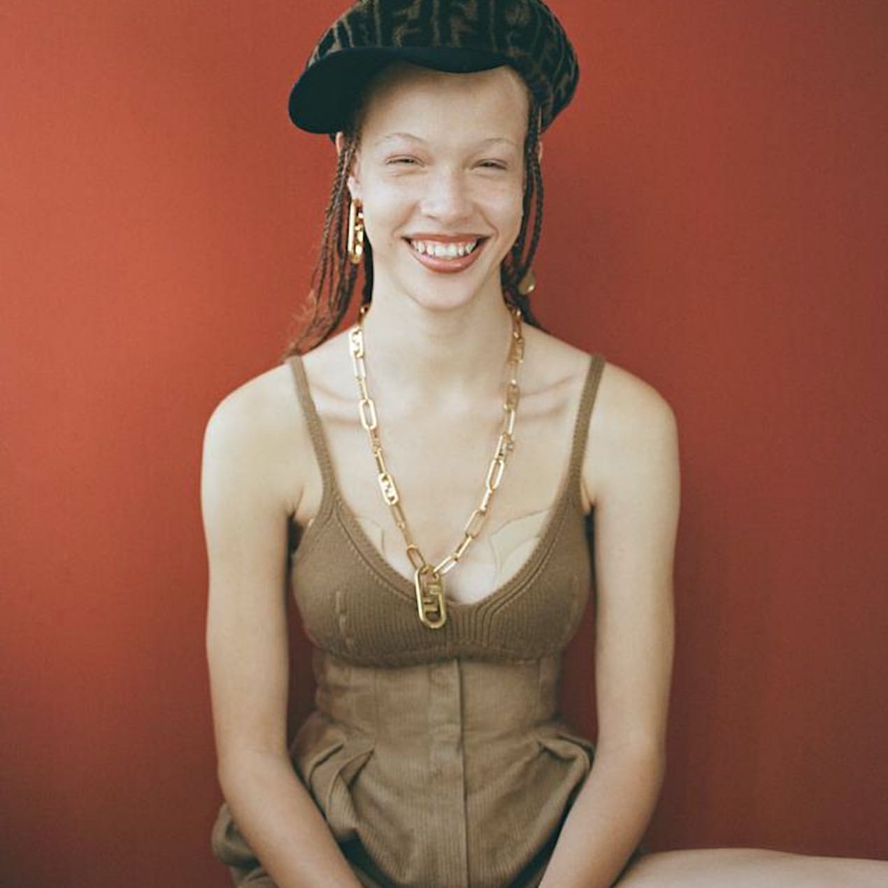 Young woman with braided hair, wearing a patterned cap, gold jewelry, and a brown dress, smiles brightly against a red background.