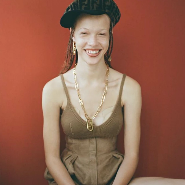 Young woman with braided hair, wearing a patterned cap, gold jewelry, and a brown dress, smiles brightly against a red background.