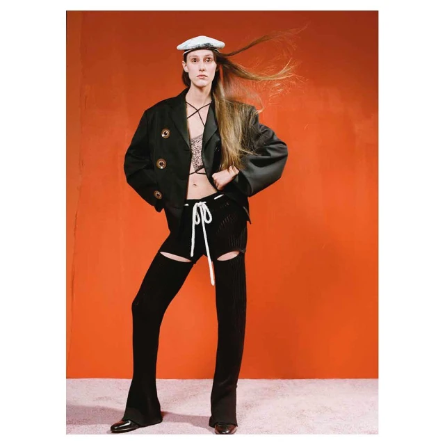 A model poses in a fashion editorial shot against an orange backdrop, wearing a black blazer, knit shorts, black stockings, and a white beret.