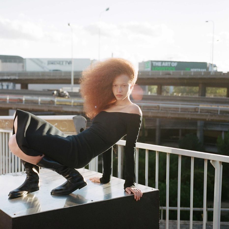Fashion shot of a red-haired woman in a black dress and boots posing on a metallic platform against an urban background.