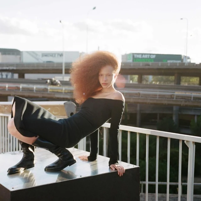 Fashion shot of a red-haired woman in a black dress and boots posing on a metallic platform against an urban background.