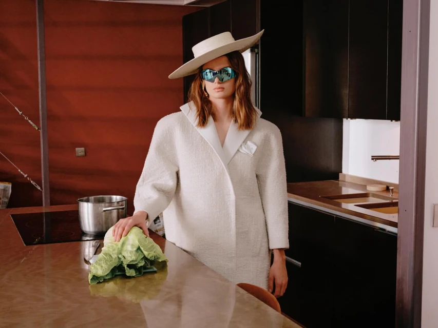Woman in a wide-brimmed hat and coat stands in a modern kitchen, holding a head of lettuce, with stainless steel appliances.
