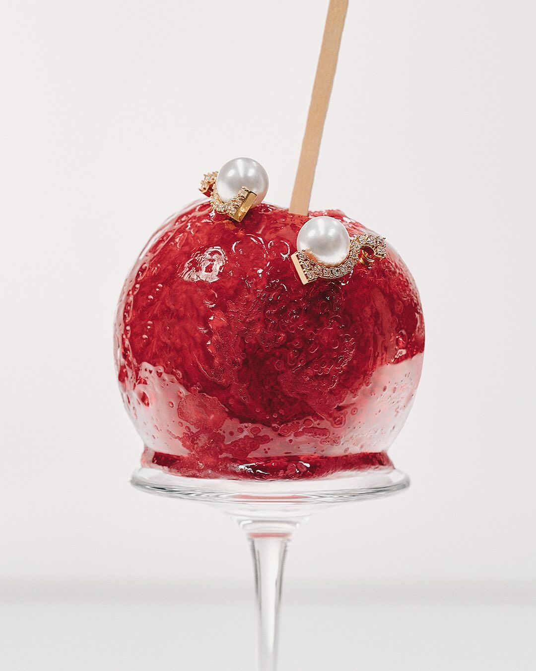 Close-up studio shot of a red candy apple adorned with jewelry, displayed on a clear glass stand against a stark white background.