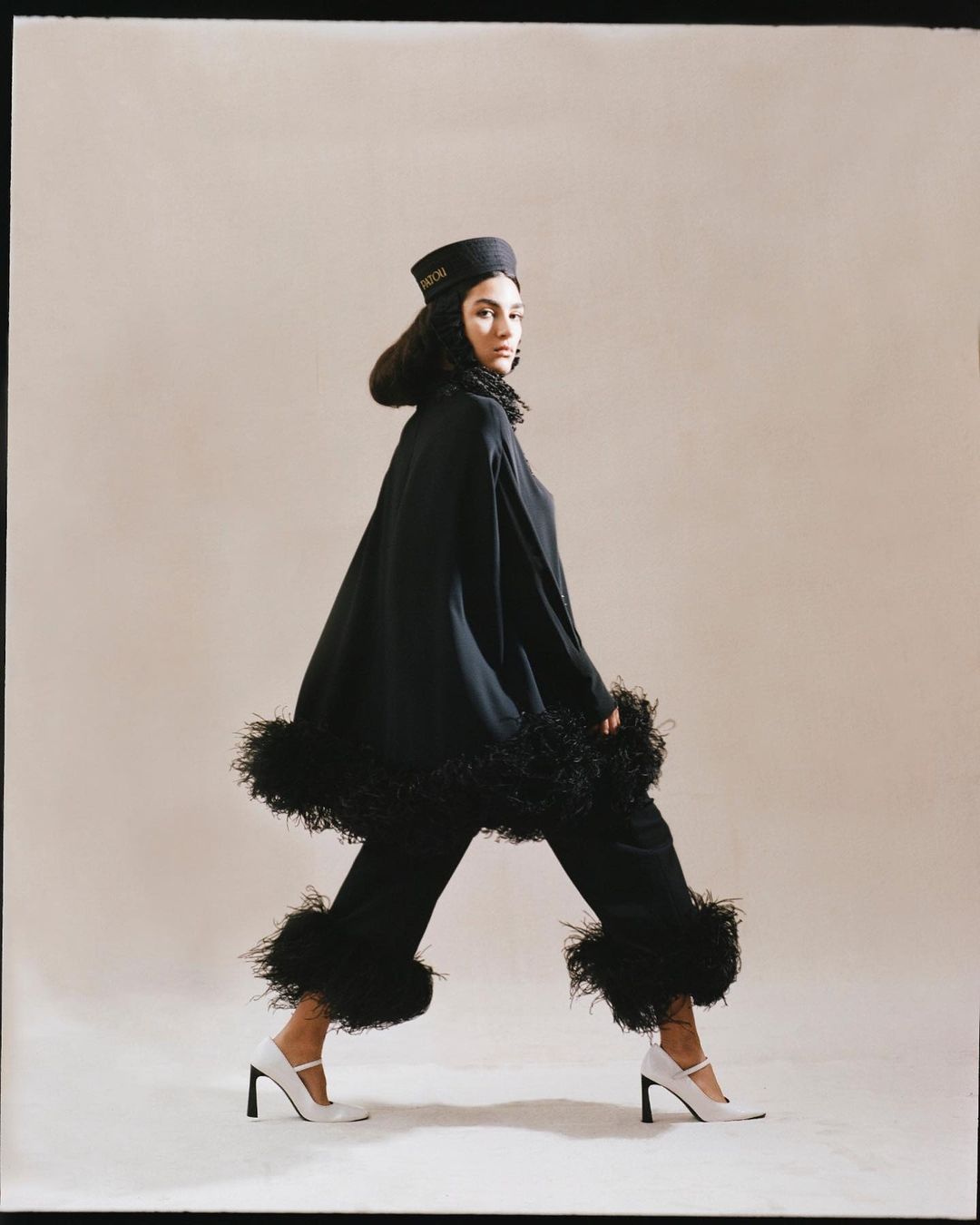 Elegant woman in a black cape and trousers with white high heels posing in a light studio with a neutral background.