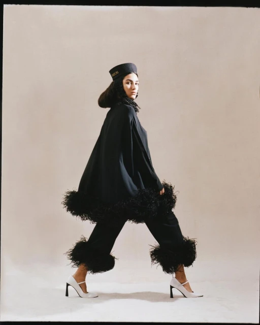 Elegant woman in a black cape and trousers with white high heels posing in a light studio with a neutral background.