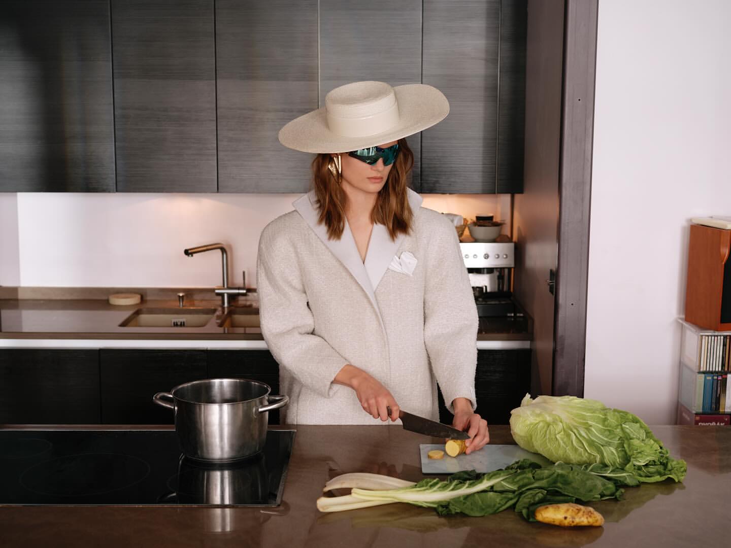 A woman in a cream-colored coat and hat prepares vegetables in a modern kitchen. She is cutting potatoes on a countertop.