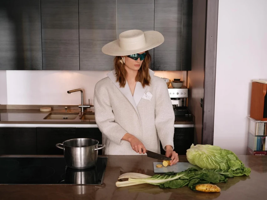 A woman in a cream-colored coat and hat prepares vegetables in a modern kitchen. She is cutting potatoes on a countertop.