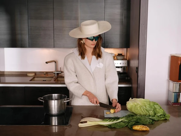 A woman in a cream-colored coat and hat prepares vegetables in a modern kitchen. She is cutting potatoes on a countertop.