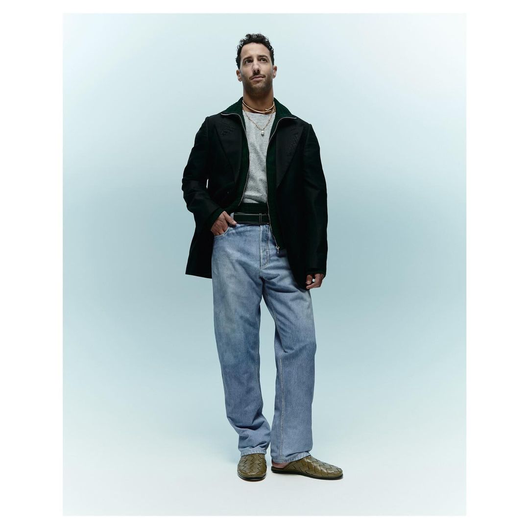 Full-length portrait of adult male wearing casual separates: dark green zip-up jacket, grey tee shirt, light wash jeans, and reptile-print shoes.
