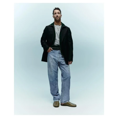 Full-length portrait of adult male wearing casual separates: dark green zip-up jacket, grey tee shirt, light wash jeans, and reptile-print shoes.