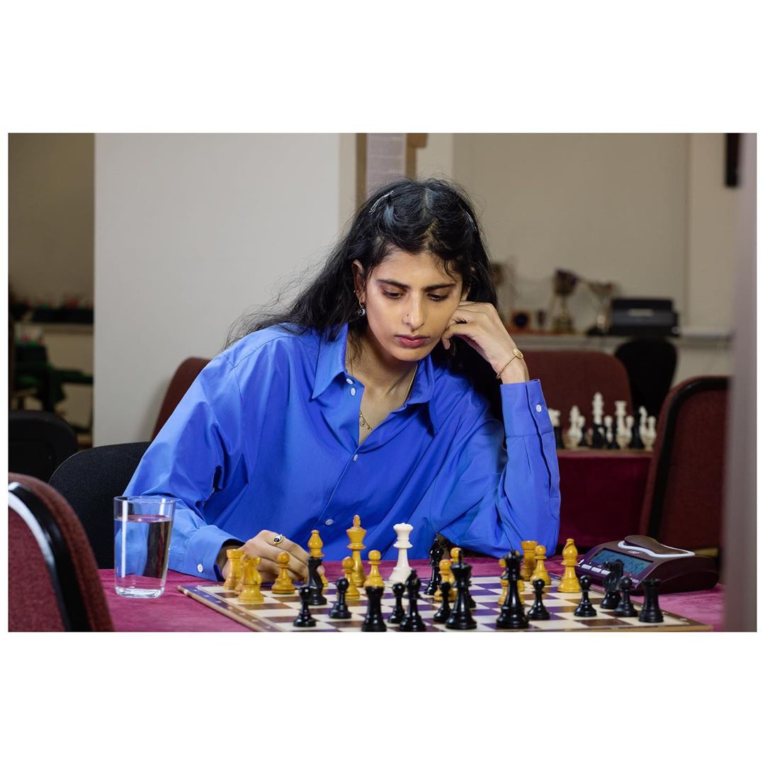 Young woman in blue shirt contemplates chess board with pieces in an indoor setting with a glass of water and clock.