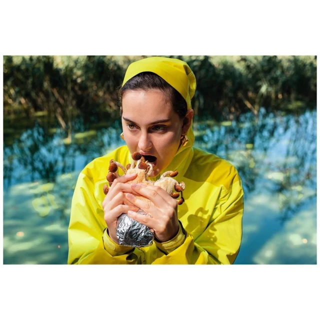 Woman in yellow eating mushrooms from a foil package in a forest setting with reflected light from a body of water.