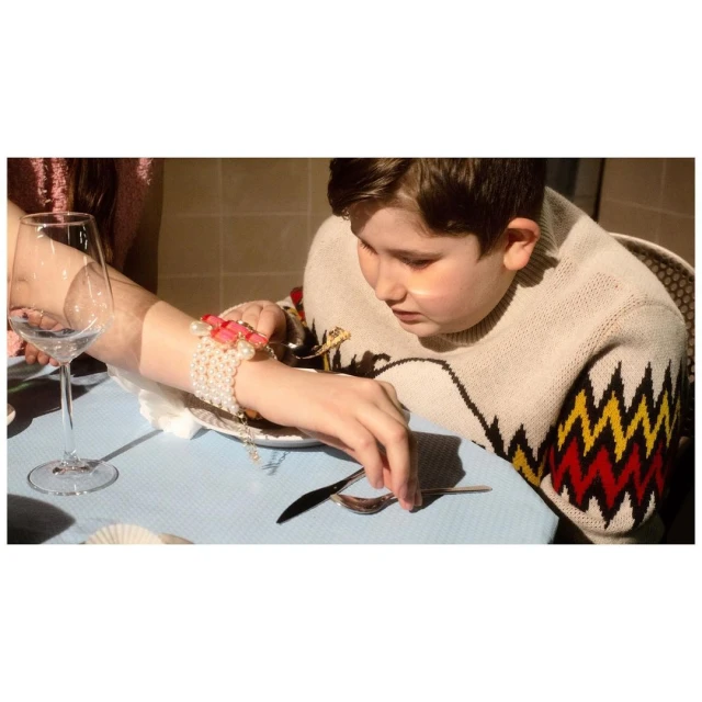 Child in patterned sweater at table set with utensils, drink, and bracelet-adorned adult arm alongside in an indoor setting.