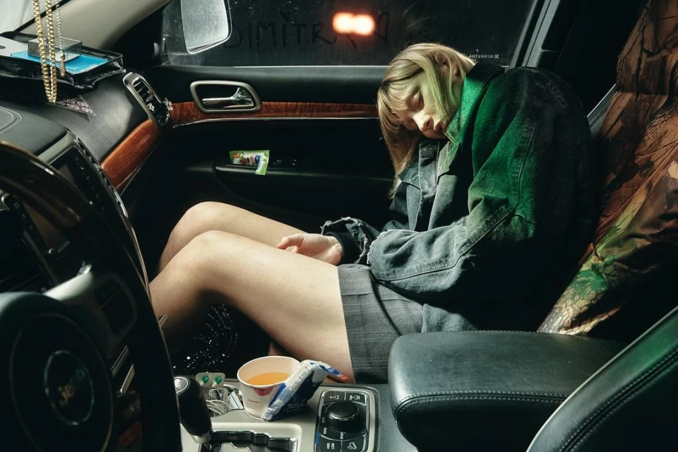 Young adult female sleeping in the driver's seat of a Jeep SUV, inside the car. She wears a denim jacket and grey skirt.