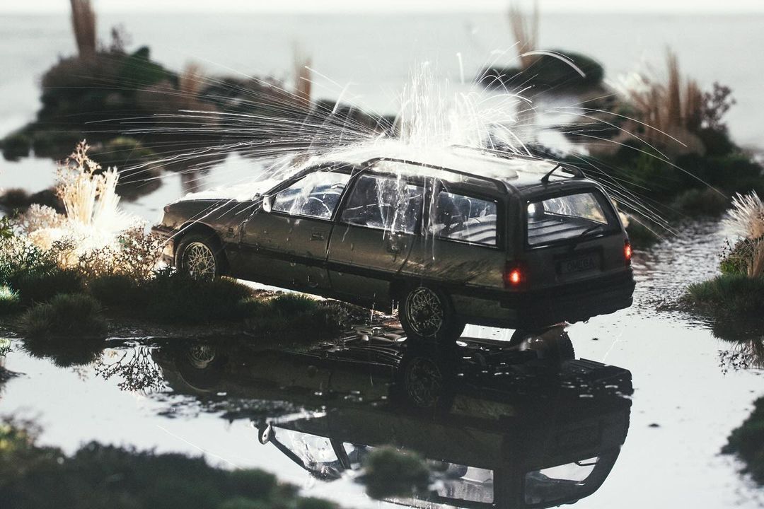 Antique station wagon vehicle is partially submerged in water as it's being struck by sparks in an outdoor setting with greenery.