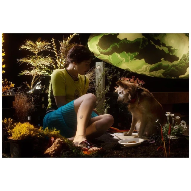 Woman sits outdoors with her dog. She wears a yellow top and blue skirt. Plants surround them; a light green background is visible.
