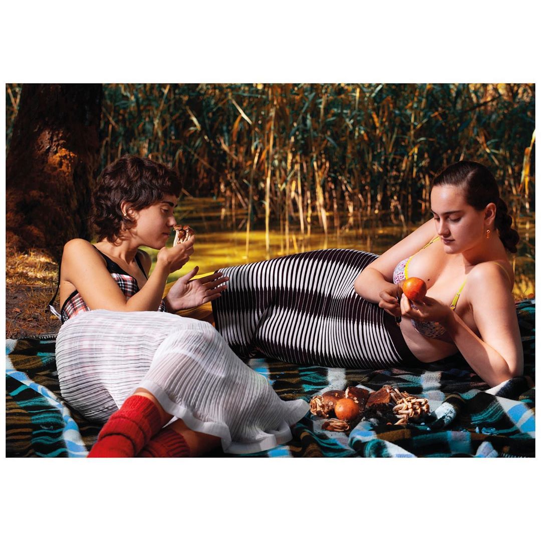 Two adult females lounge on a patterned blanket near the water's edge surrounded by tall grass. One holds fruit, picnic food is nearby.