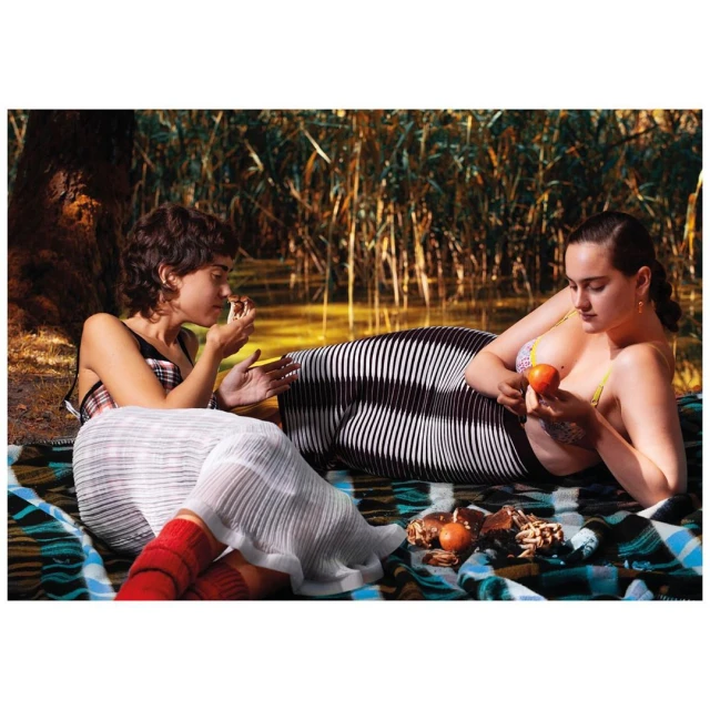 Two adult females lounge on a patterned blanket near the water's edge surrounded by tall grass. One holds fruit, picnic food is nearby.