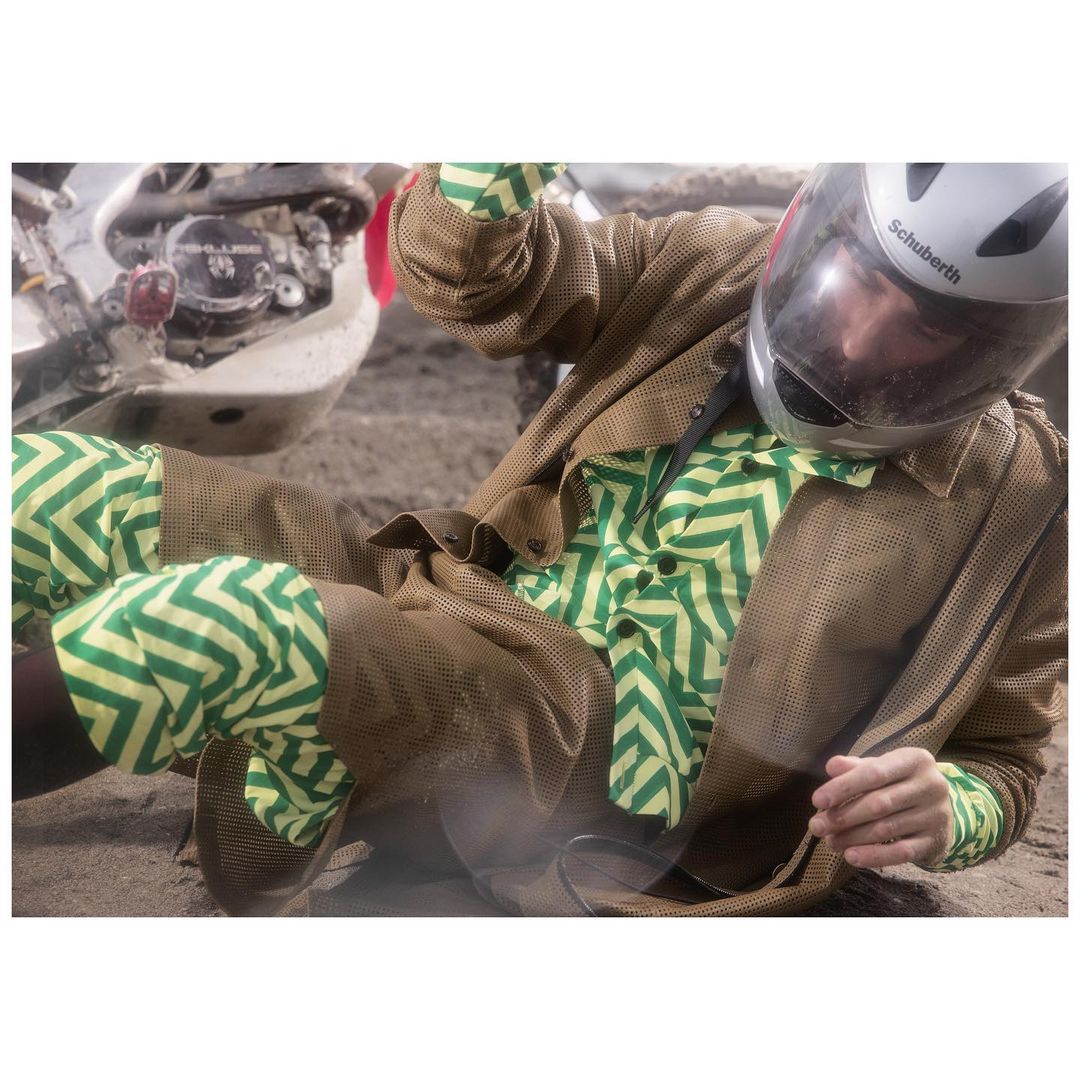 Man in helmet and patterned shirt lying on ground near motorcycle. Fashion shot with gritty sand environment. Automotive, active lifestyle.