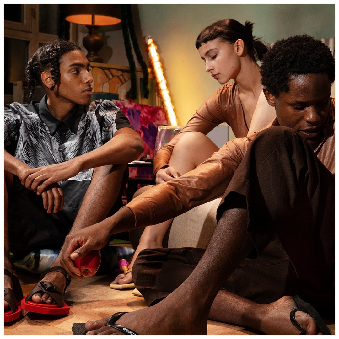 Three diverse adults are seated on the floor in an indoor setting, wearing fashionable clothing and sandals, with colorful decor behind them.