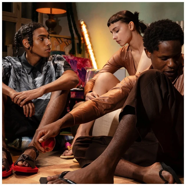 Three diverse adults are seated on the floor in an indoor setting, wearing fashionable clothing and sandals, with colorful decor behind them.