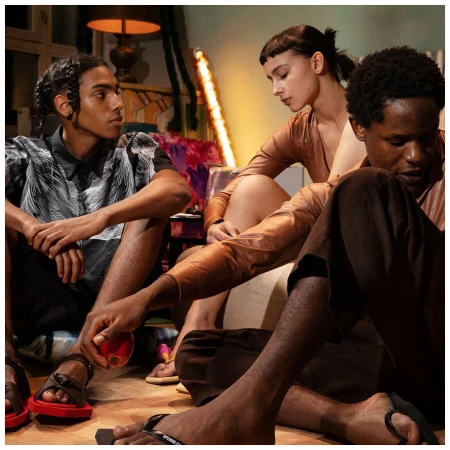 Three diverse adults are seated on the floor in an indoor setting, wearing fashionable clothing and sandals, with colorful decor behind them.