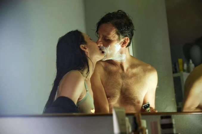 A couple is seen in a bathroom. A brunette woman kisses a man who has shaving cream on his face while holding a razor.