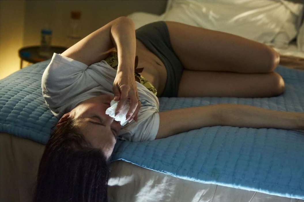 Adult woman lying on a blue bed with white linens, holding a tissue to her face, indoors in a dimly lit room with a side table.